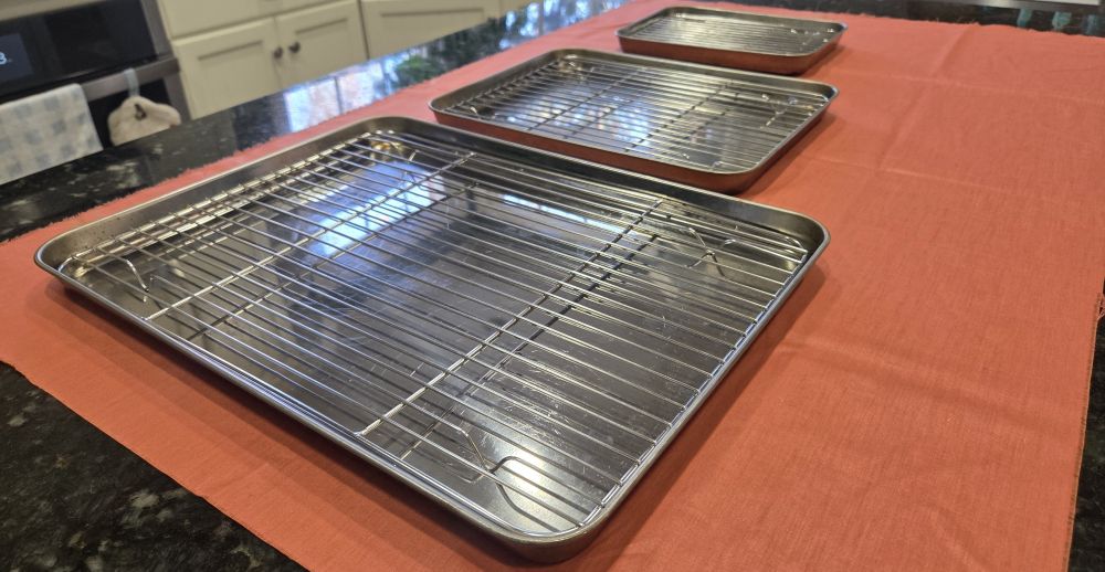 Three stainless steel baking sheets of descending sizes, and accompanying cooling racks, are arrayed across a coral-coloured fabric swath on a countertop. Household products for baking, like these sheets, should ideally be made of non-toxic materials to reduce exposure to toxins in the food we eat and serve others.