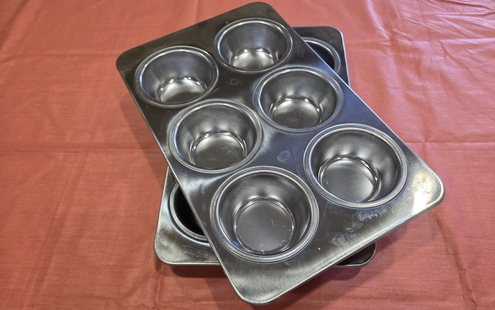 Top-down view of two 6-cup stainless steel muffin pans, one stacked askew atop the other. These are useful household products to have for toxin-free cupcakes and muffins