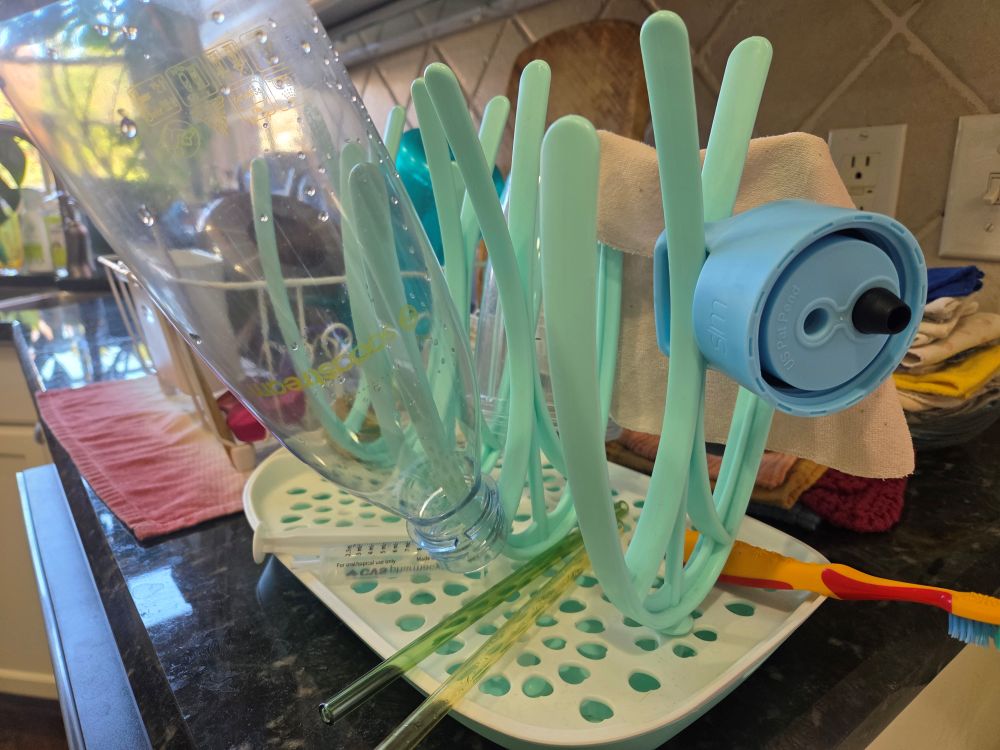 A baby bottle drying rack, loaded with various smaller dish items such as a medicine syringe, toothbrush, glass straws, water bottle parts, etc. sits atop a counter next to the normal dish drying rack