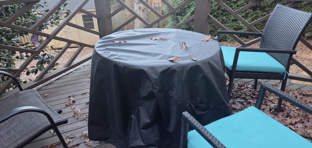 Black waterproof round patio table cover draped over a deck table with a few chairs around and fallen leaves nearby and on top