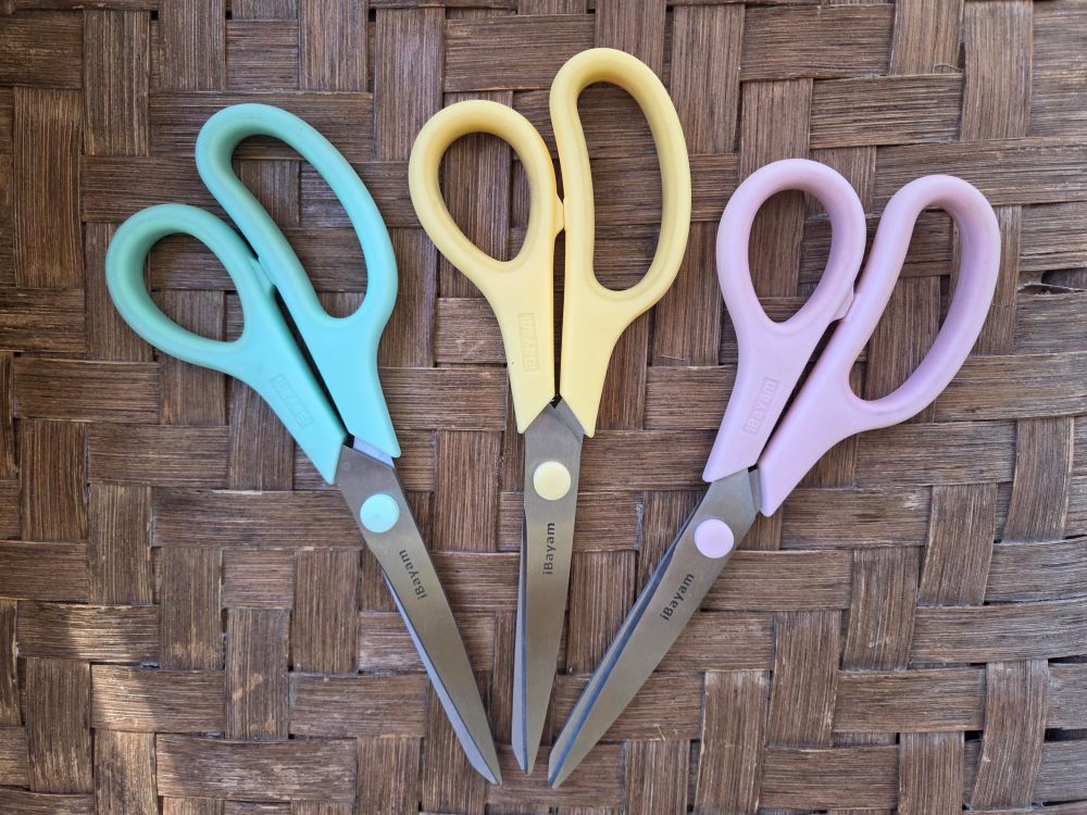 Three pairs of pastel kitchen scissors arrayed on the seat of a woven/wicker chair, in sea green, pale yellow and pale pink. If you can find pretty versions of common household products like scissors, opt for those!