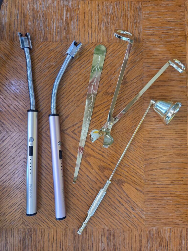 Two electric lighters in silver and rose gold and a gold-coloured metal candle care set (consisting of snuffer, wick trimmer and wick dipper) viewed top down on a wood table