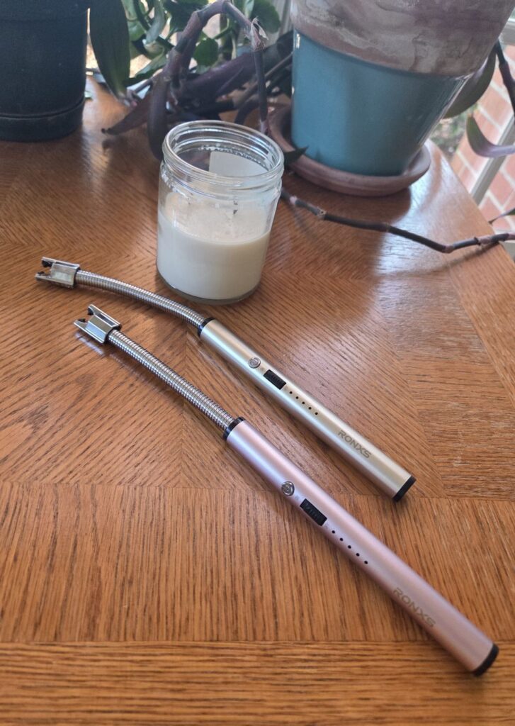 Two electric lighters in silver and rose gold lay atop a wood table nearby a candle and some plants