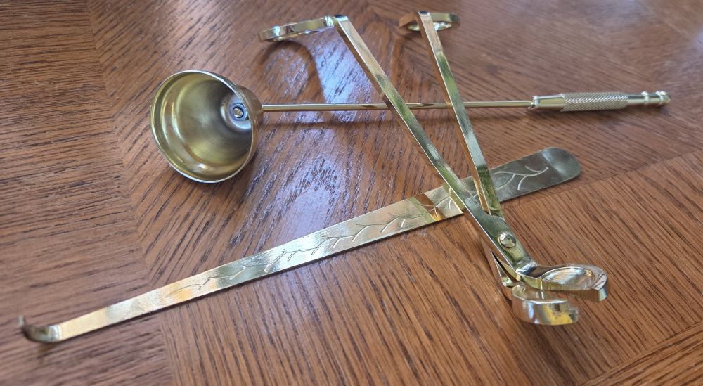 A trio of gold-coloured metal candle care items consisting of bell-shaped snuffer, scissors-like wick trimmer, and wick dipper rest on a wood table