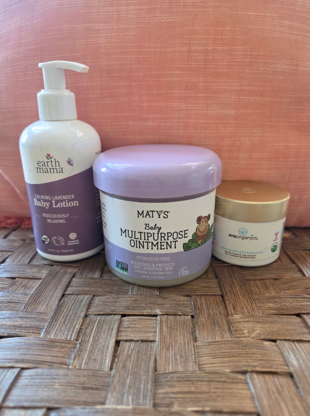 A bottle of lavender baby body lotion, a jar of Maty's multipurpose ointment, and a jar of Era Organics balm rest in front of a coral-coloured pillow on the seat of a wicker chair