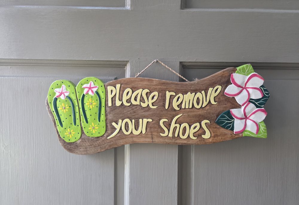 A carved/painted wooden sign hangs on an exterior door and reads 'Please remove your shoes' next to a carved/painted picture of decorative flip flops