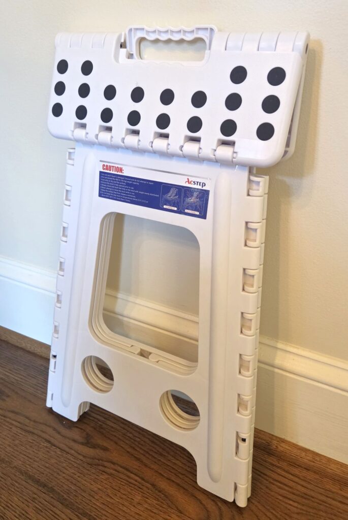 A white stepstool with grippy black circles/dots design rests folded up against a wall