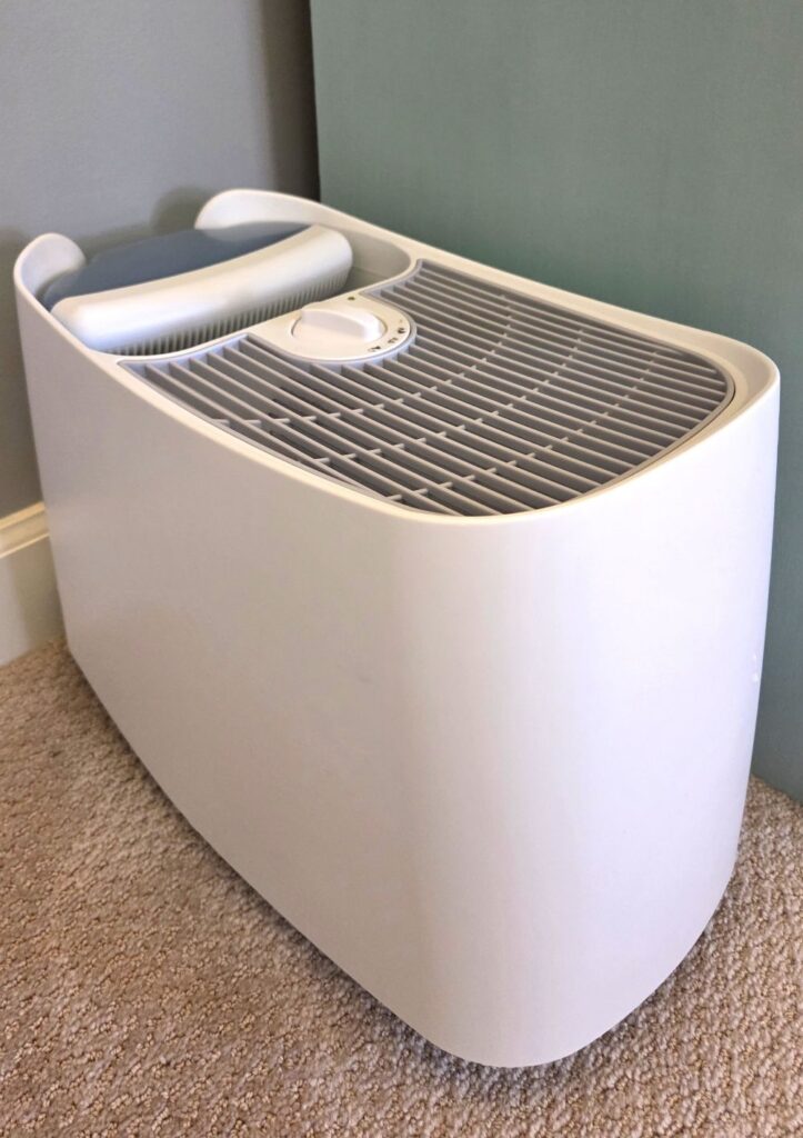 A white cool-mist humidifier with its water tank sits on the floor of a carpeted bedroom next to a dresser