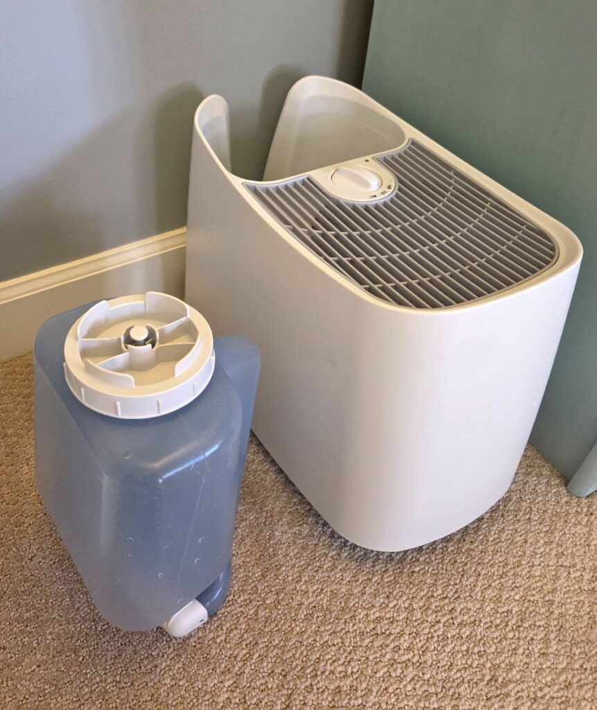 Picture of a white cool-mist humidifier with its water tank removed and sitting beside it on the floor of a carpeted bedroom next to a dresser