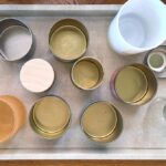 Top-down view of an off-white faux leather rectangular tray with silver-coloured metal handles on either end resting atop a wood table and filled with a number of small jars of various materials, sizes and colours