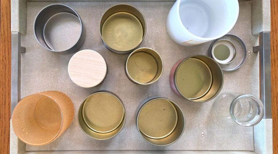Top-down view of an off-white faux leather rectangular tray with silver-coloured metal handles on either end resting atop a wood table and filled with a number of small jars of various materials, sizes and colours