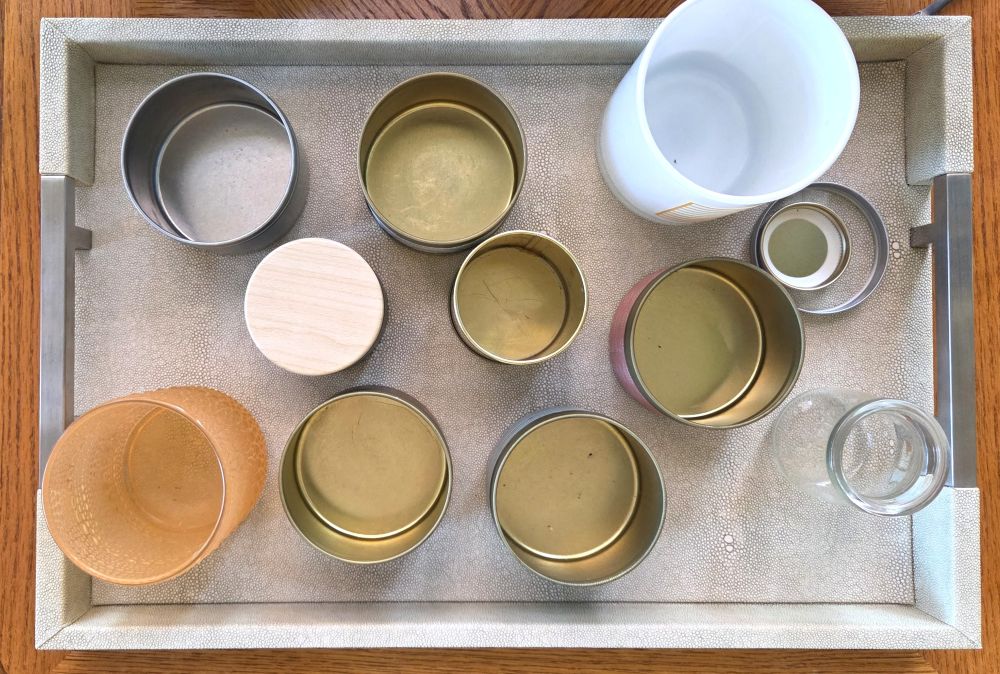 Top-down view of an off-white faux leather rectangular tray with silver-coloured metal handles on either end resting atop a wood table and filled with a number of small jars of various materials, sizes and colours