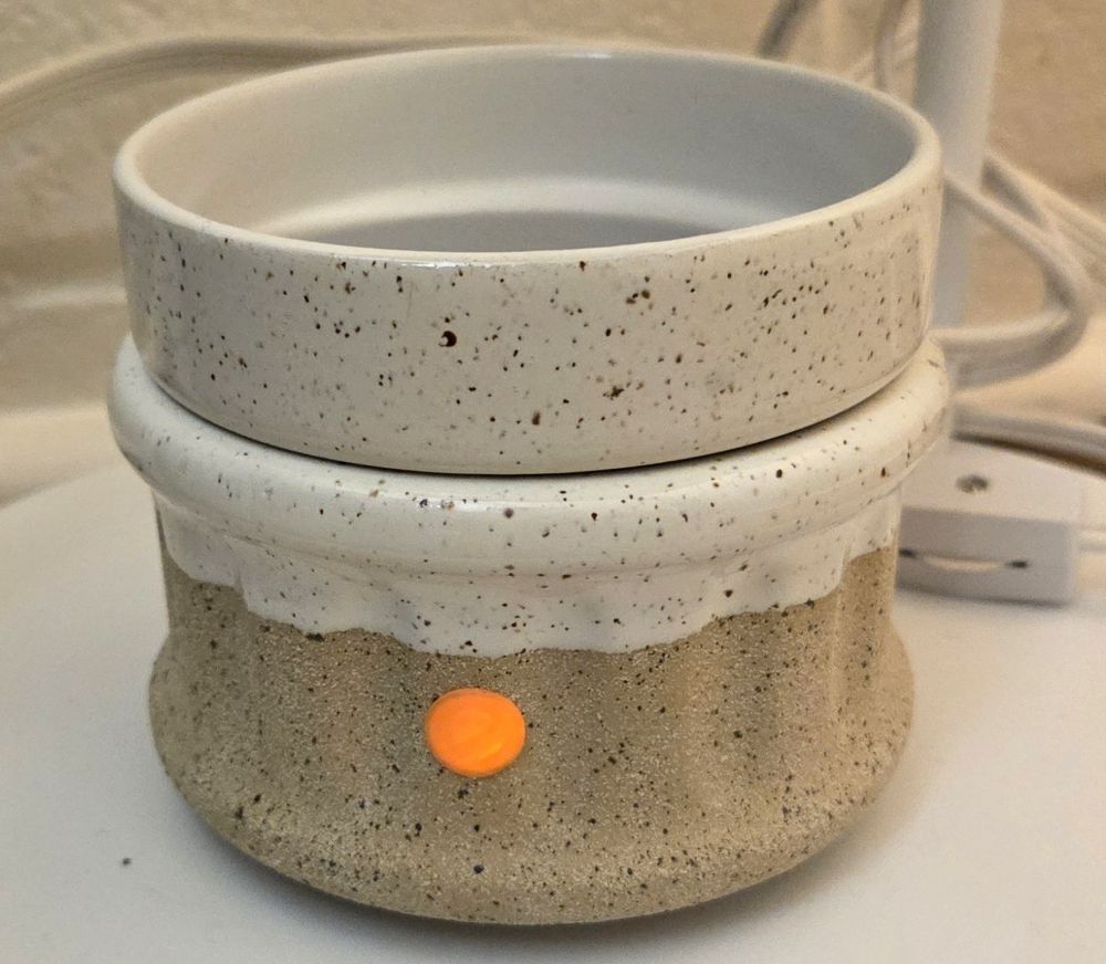Close-up image of a ceramic wax melt warmer and the switch on the cord wrapped around a table leg behind it, with the small orange light on the front lit up to indicate it's turned on