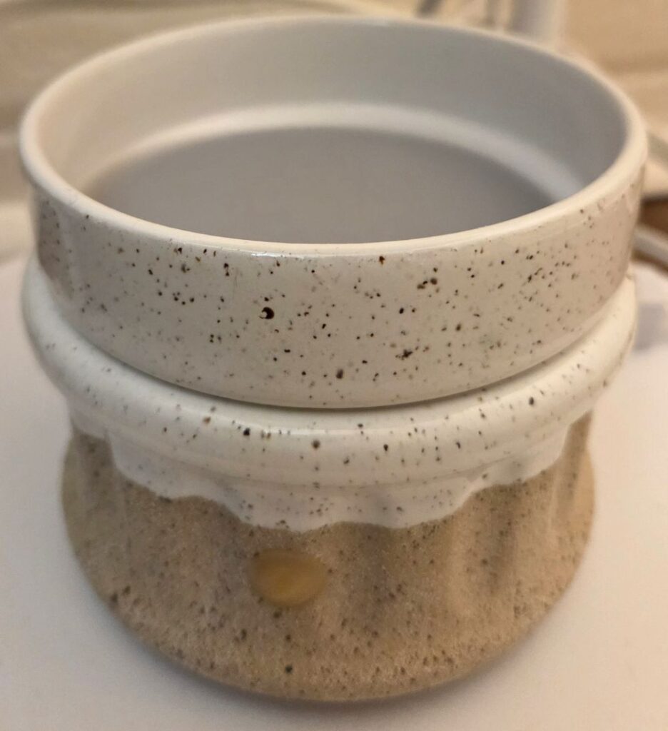 Close-up image of a natural-looking ceramic wax melt warmer in a sand-coloured hue with creamy white paint around the top, and the switch on the cord wrapped around a table leg behind it