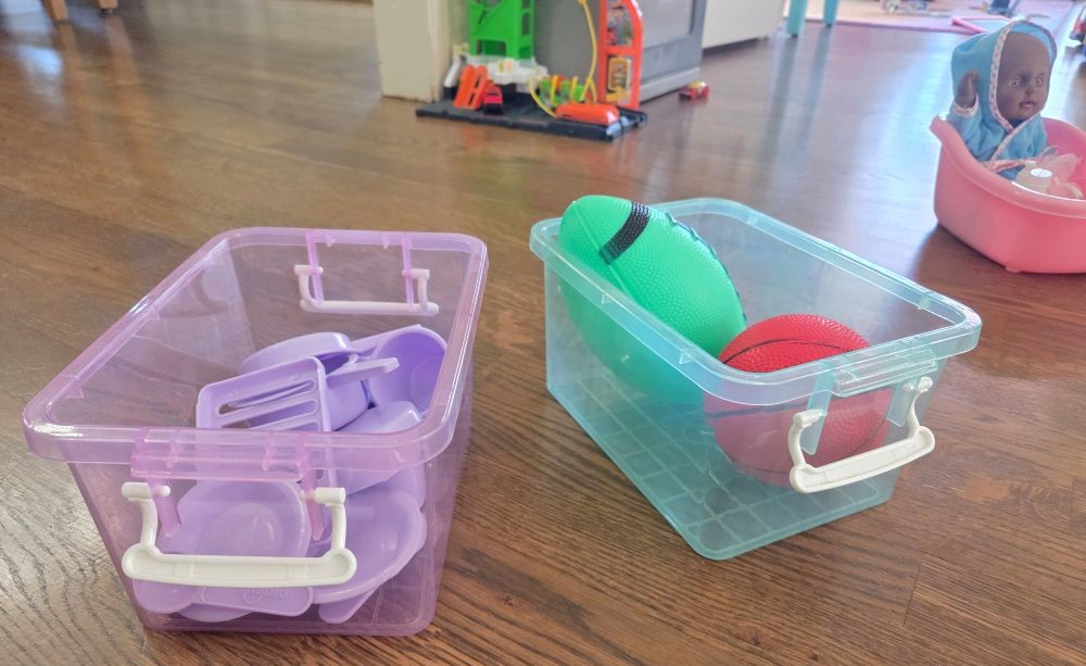 Two clear pastel-coloured rectangular storage bins (in lavender and sea green) are filled with small toys and sitting on a wood floor. If you are drawn to certain colours, you can find common household products (like storage bins) in those colours!
