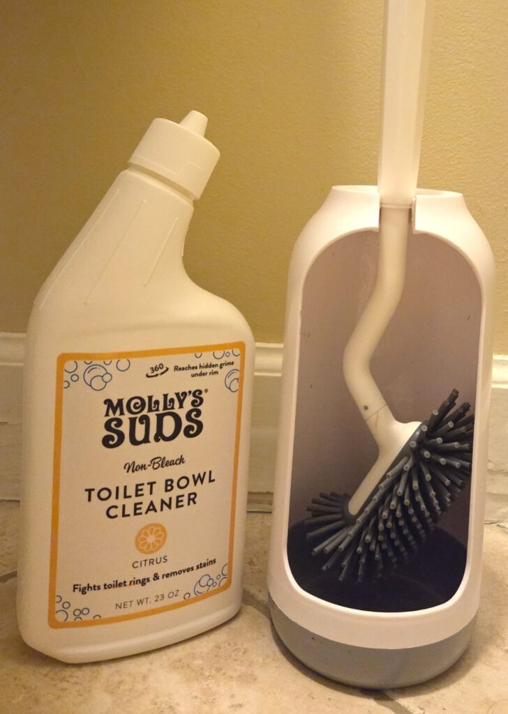 Bottle of Molly's Suds toilet bowl cleaner in corner of bathroom next to silicone 'bristled' toilet brush hanging in touch-free stand