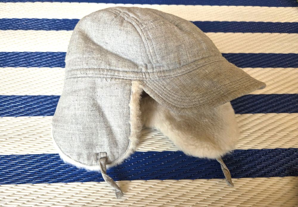 Atop a blue and white striped rug rests a light gray toddler-sized white faux-fur-lined winter hat with a short 'bill' in front for sun shade and two short strings on either side of ear overhangs