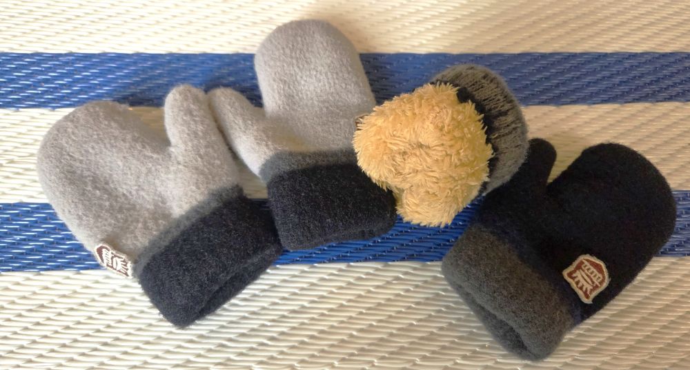 Atop a blue and white striped rug rest two pairs of fleece-lined toddler-sized mittens spread in an array in navy and gray. One of the mittens is shown turned partially inside-out to reveal the thick insulated interior