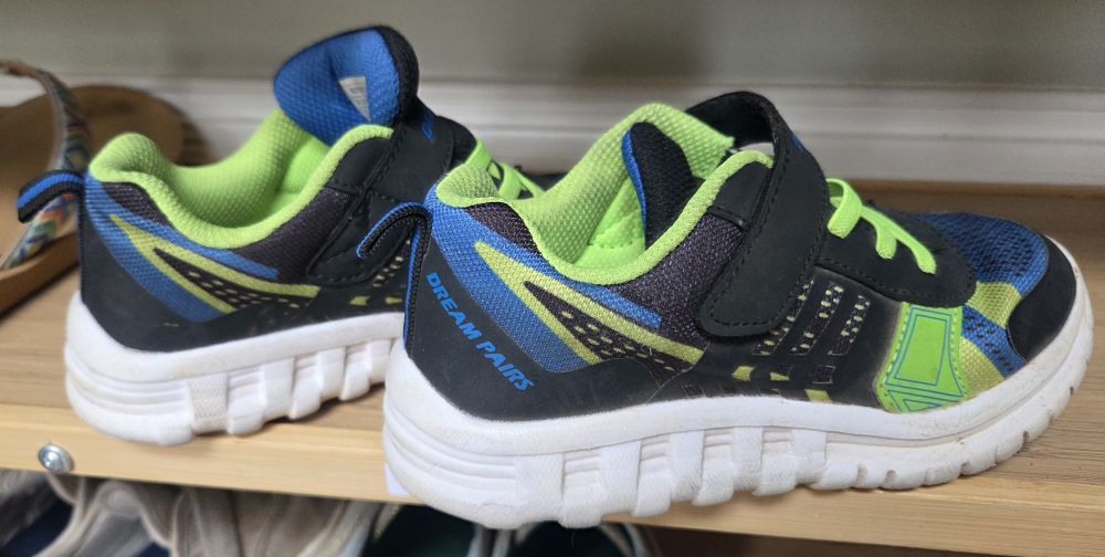 Close-up view of a pair of children's Dream Pairs athletic sneakers with Velcro straps (and non-functional/decorative laces) in black, electric blue, and neon lime detailing. The white bottom of shoe is thick and ergonomically-shaped