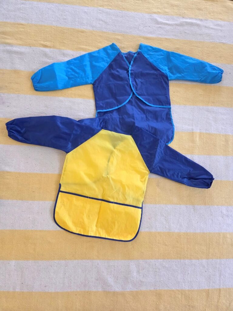 Two long-sleeved colourful toddler/children's painting smocks, in blue and yellow, are spread out atop a yellow and white striped rug, with the blue one turned over to see the back side which ties shut like an apron