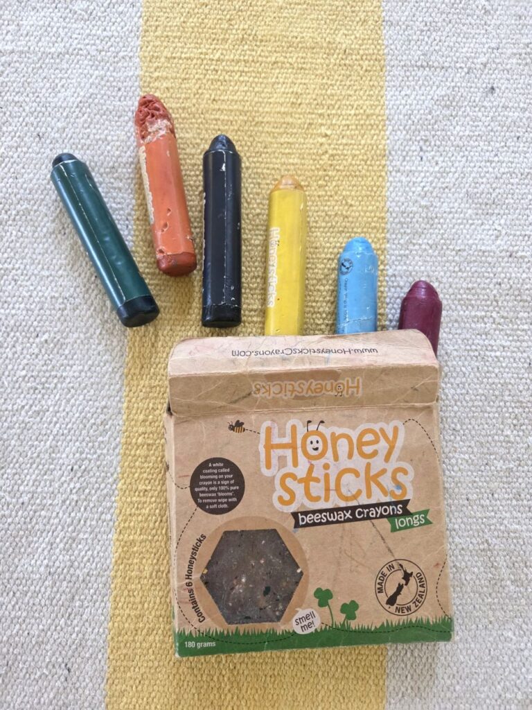 A pack of Honeysticks beeswax baby crayons lays open atop a yellow and white striped rug with all six large-sized crayons (in green, orange, black yellow, blue and red) sticking out of the pack
