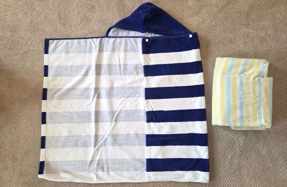 Two striped hooded toddler towels lay on a carpeted floor. The dark blue and white striped one has part of it folded inside and part of it open to show both the outside and inside, as well as the white snaps at the top. The light blue, white and yellow striped one (exact same design) sits folded beside the other one.