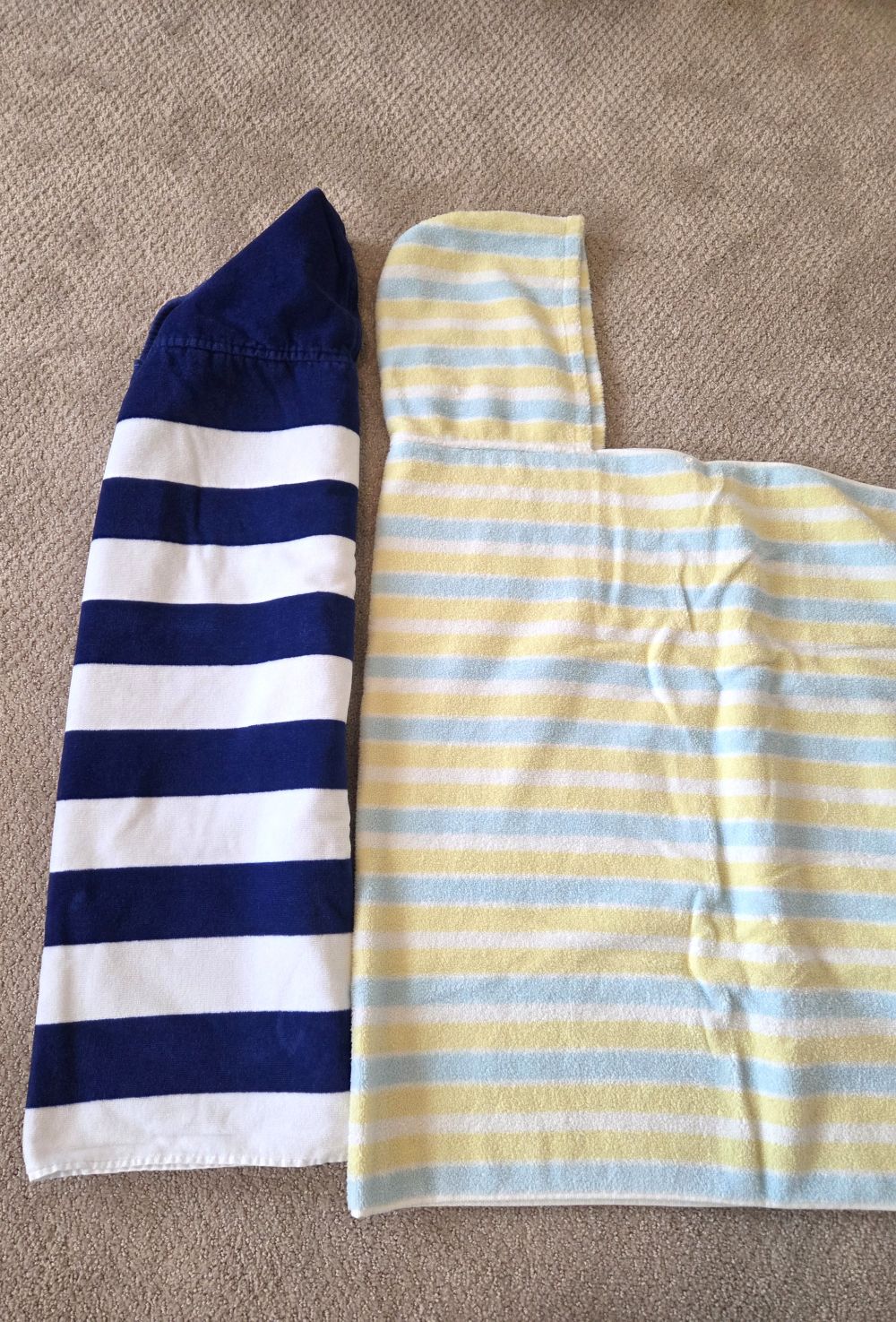 Two hooded toddler towels lay on a carpeted floor. The dark blue and white striped one is extended lengthwise and tucked inside width-wise. The light blue, white and yellow striped one (same shape/design) is also extended lengthwise but tucked in just once widthwise to show the hood and one side extended