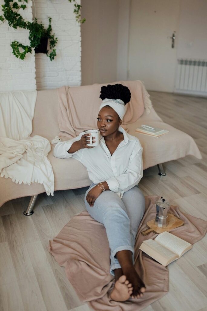A woman rests over a blanket on the floor while leaning against a sofa covered in blankets and drinking a mug of coffee next to an open book and carafe