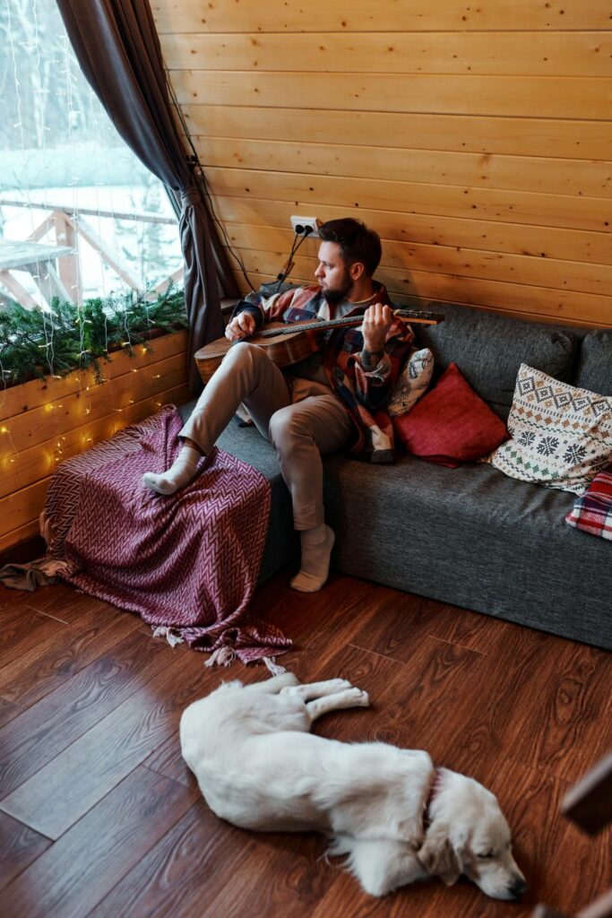 A man sits atop a couch with pillows and a blanket in the corner of wood-paneled A-frame house by the window playing guitar while a dog rests on the wood floor nearby