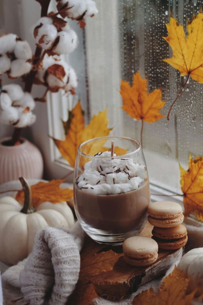 An unlit candle that resembles a hot cocoa with whipped cream and chocolate sprinkles in a clear glass rests next to macarons in front of a rainy window next to a pumpkin and other natural furnishings