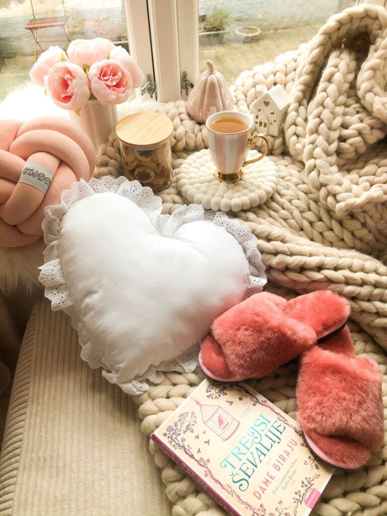 A soft woven cream-coloured wool blanket is folded about a cream-coloured corduroy suede sofa with a pair of pink slippers on top, a book, a white heart-shaped pillow, a pink knot pillow, a candle and flowers