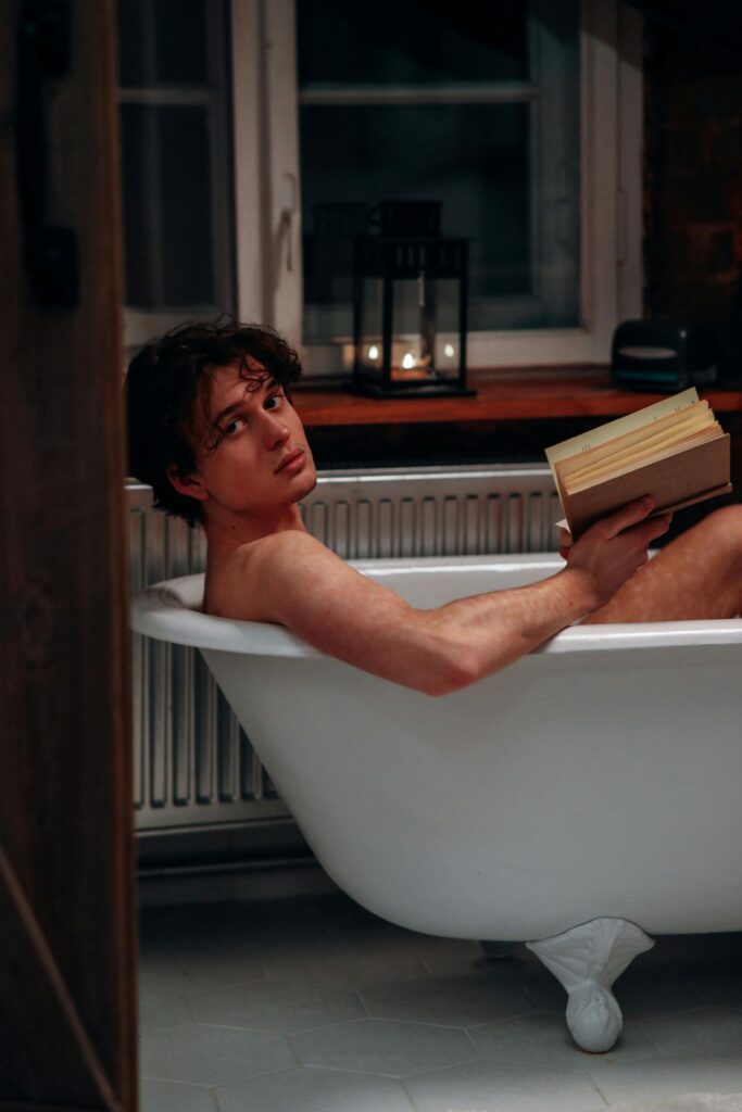 A man relaxes in a clawfoot bathtub while reading a book next to a lit candle on the windowsill