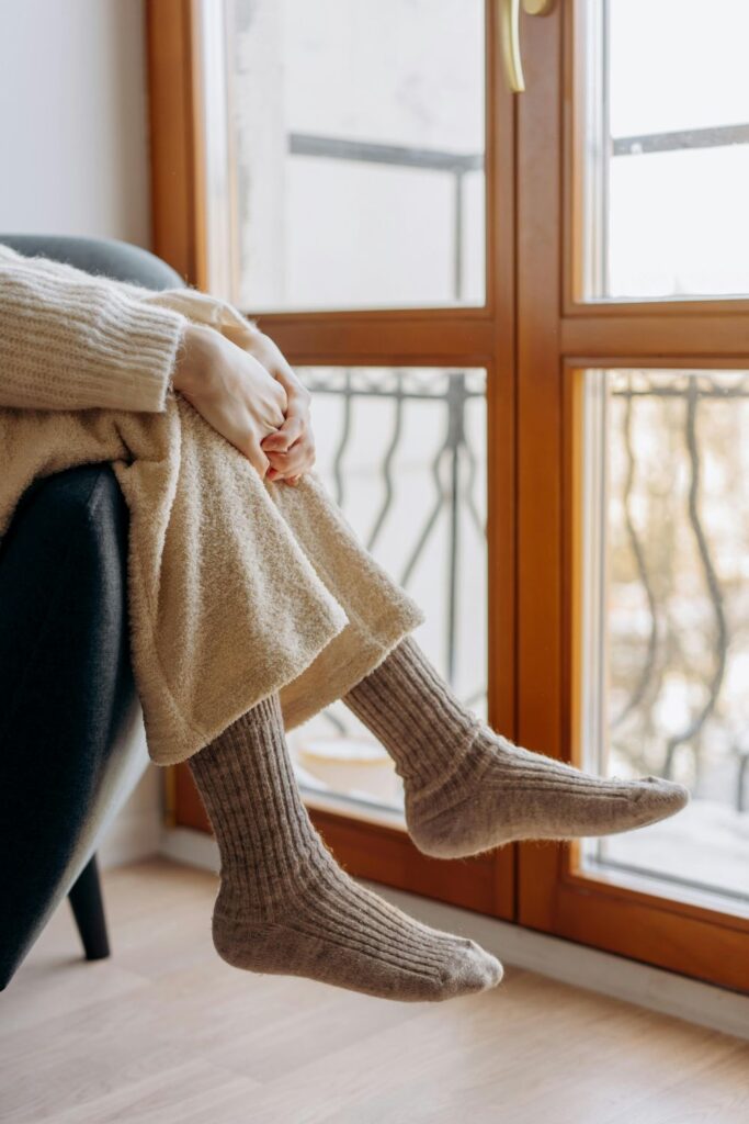 A woman wearing soft pants, sweater and cosy socks lounges over a sofa armrest in front of a balcony door/window
