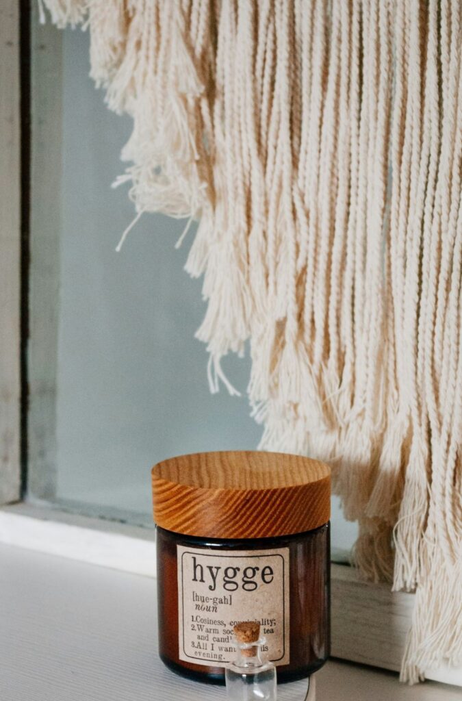 A scented candle titled 'hygge' sits on a windowsill in front of a tapered macrame curtain