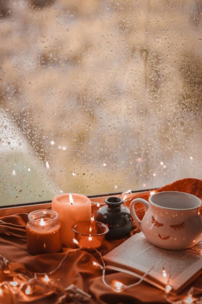 A few small lit candles, a mug, string lights, and an open book sit atop a blanket on a windowsill in front of rainy window