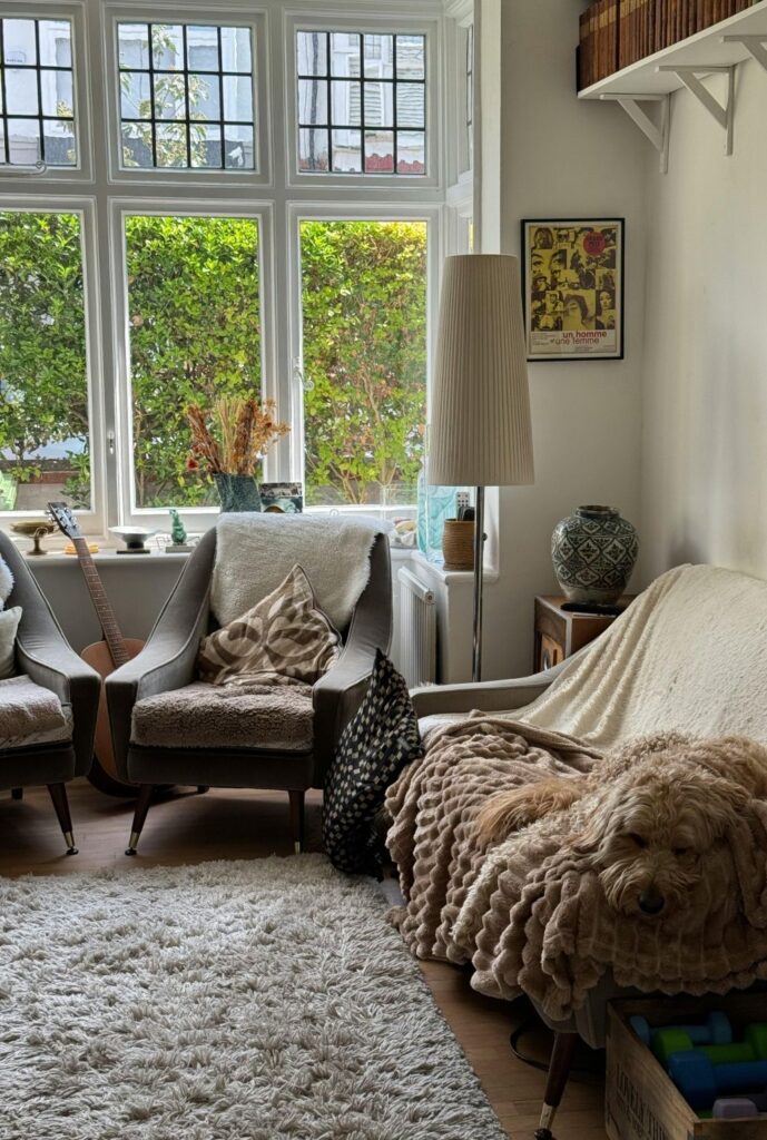 The corner of a cosy living area is furnished with a soft rug and upholstered chairs and a loveseat covered in blankets and pillows in natural colours