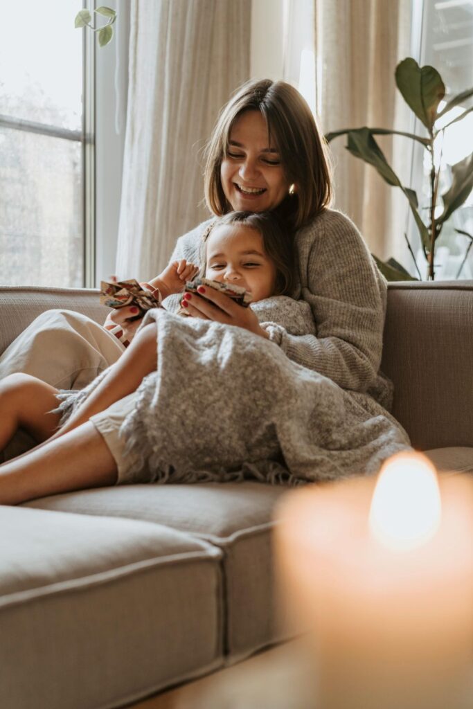 A woman and her young daughter snuggle atop a sofa in the corner by windows and a plant with a blanket and a lit candle seen in the foreground on the coffee table