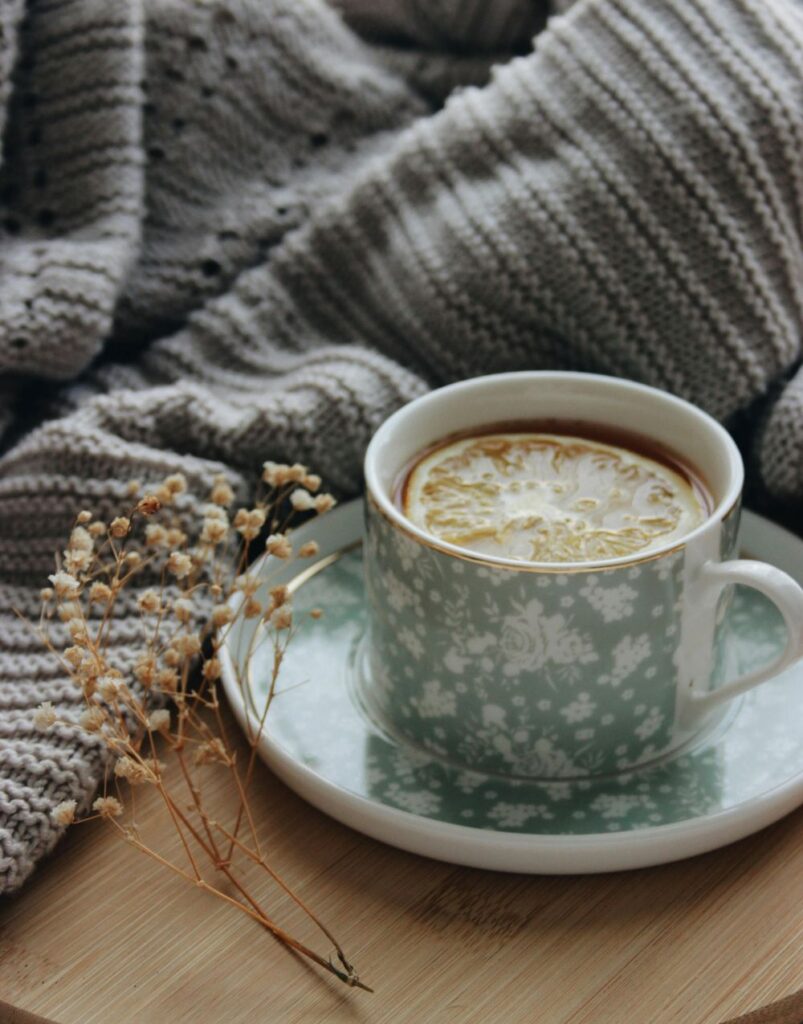 A mug of tisane with an orange slice floating inside rests on a small wooden table next to a soft knit gray sweater