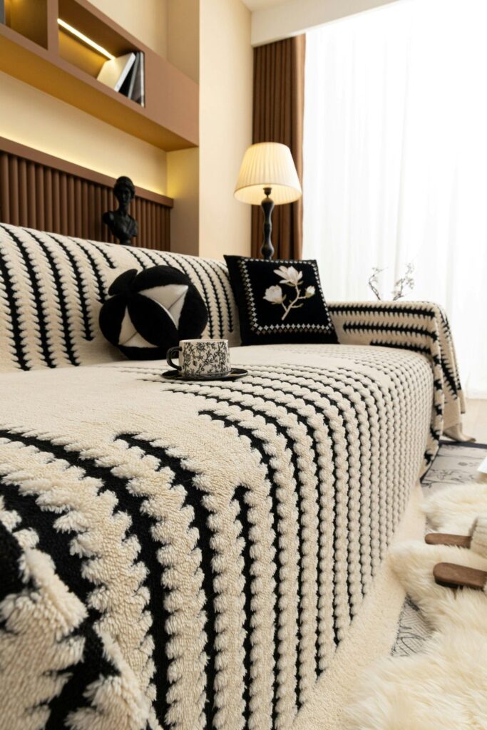 A sofa with a soft black and white slipcover is seen next to a soft white rug, a pair of slippers, and black and white throw pillows (as well as a black and white mug of coffee) on top