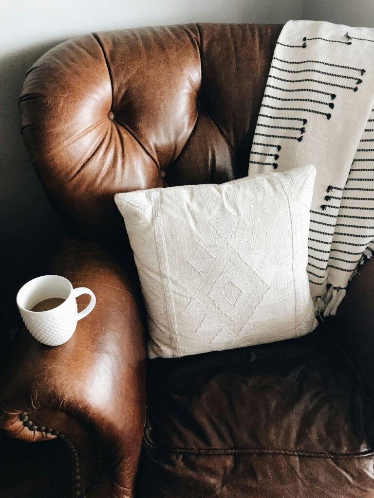 A brown leather armchair is seen with a black and white woven blanket folded over top, a white throw pillows, and a white mug of coffee resting on the armrest