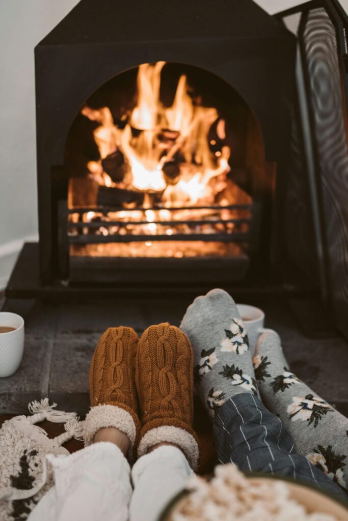 Two pairs of feet are seen relaxed in front of a lit fireplace, one clad in warm slippers and the other in cosy socks