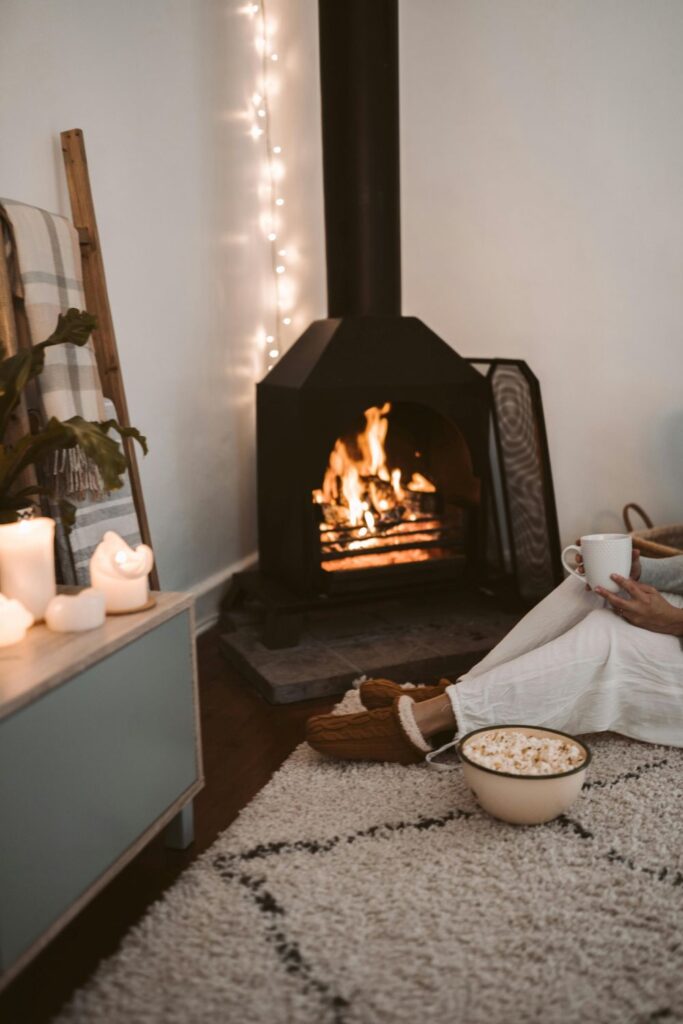 A person wearing slippers sits in front of a lit corner stove atop a soft rug, holding a mug with a bowl of popcorn, and lit candles on a low table nearby