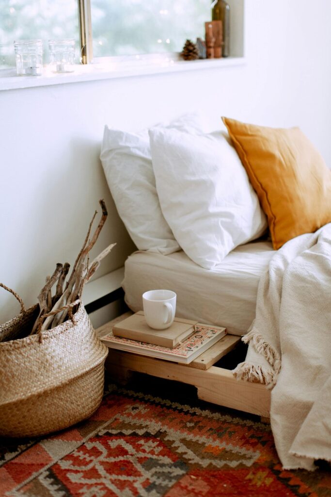 A low-to-floor bed with mattress, blanket and cushions is seen with a mug and books next to it along with woven rug and woven basket filled with sticks for a calm and naturally furnished bedroom