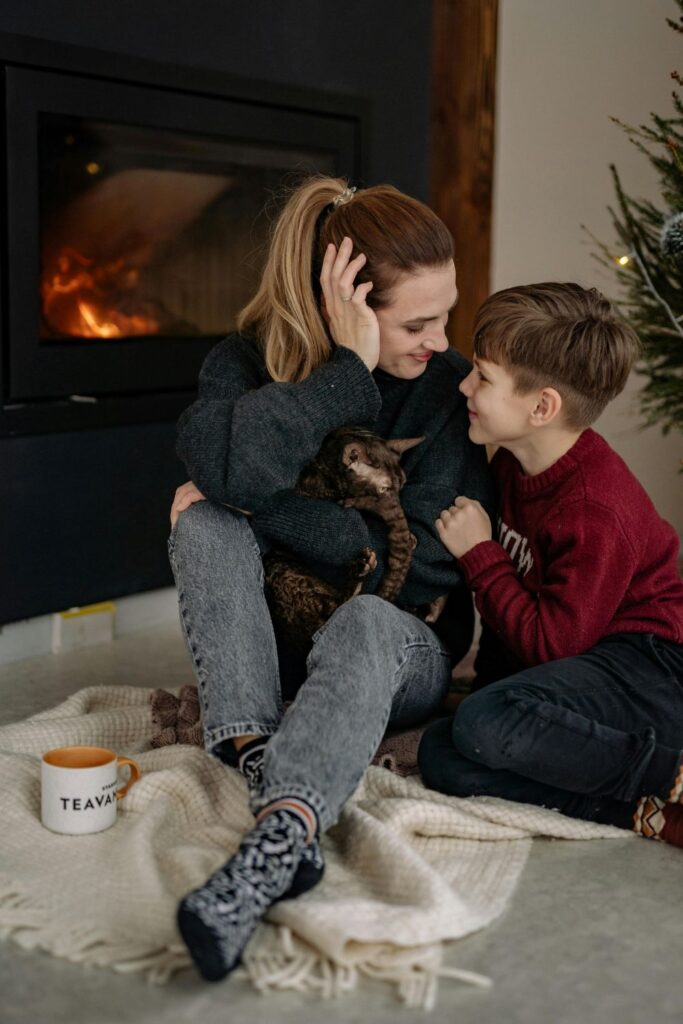 A woman holds a cat and sits cosily on a rug in front of a lit fireplace next to her son and a mug