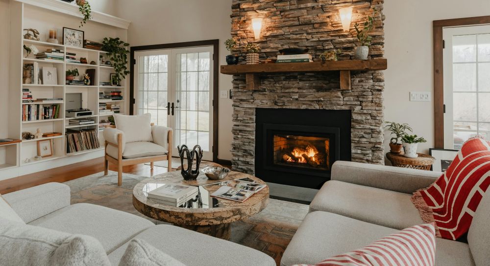 A large living space in Rhinebeck, New York features lots of natural light as well as softly-lit wall sconces above a stone fireplace with lit fire. A large light-coloured fabric sofa surrounds a round wooden coffee table and is covered in a blanket and cushions. Across the coffee table is a matching chair atop a large area rug and nearby shelves with plants and books