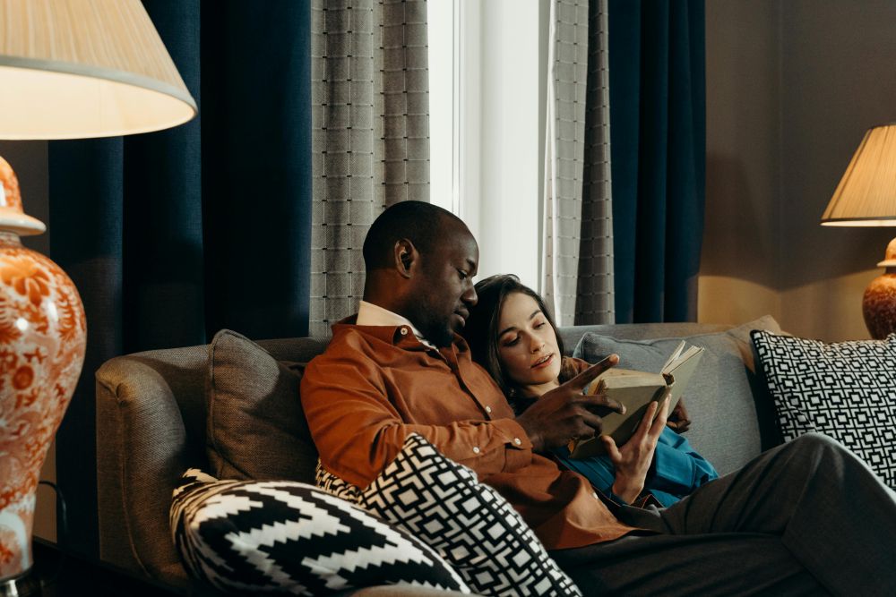 A couple smuggles on a sofa with cushions and softly-lit lamps nearby while reading a book together