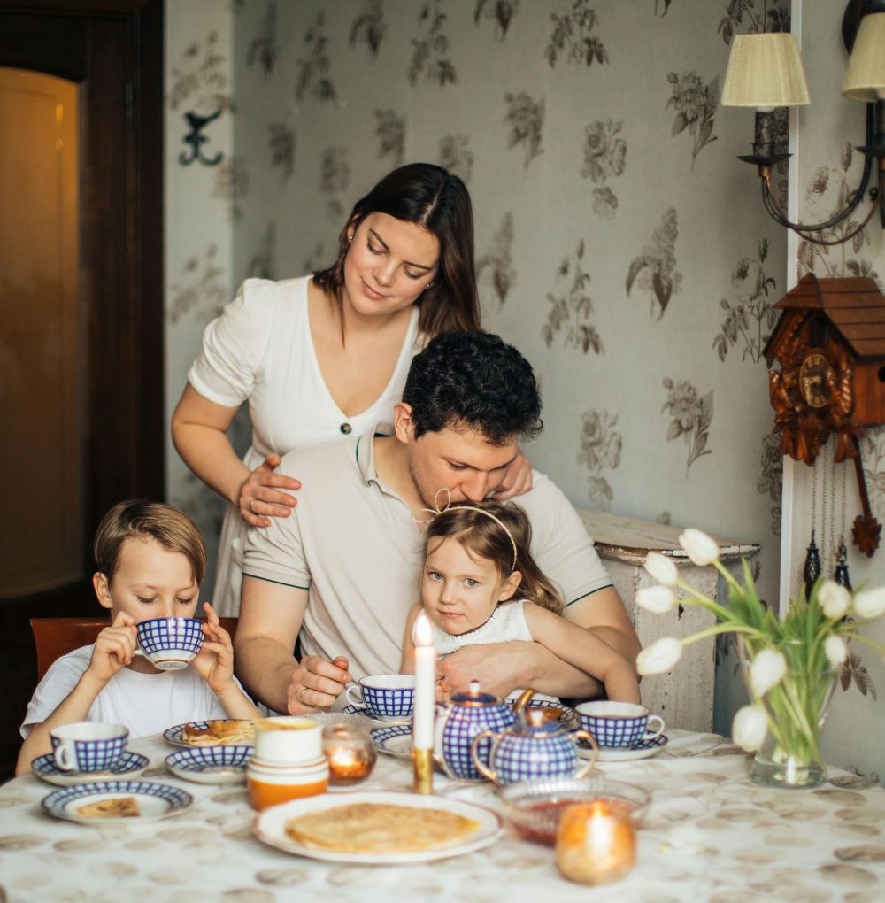 A couple and two children sit at the kitchen table eating blini with tea in blue and white chequered china with a lit taper candle and natural floral furnishings