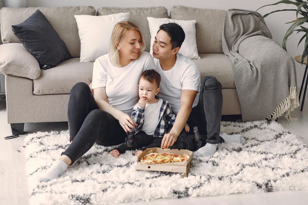 A couple and their toddler relax on a soft high pile rug in front of a sofa with a blanket and cushions while eating a pizza