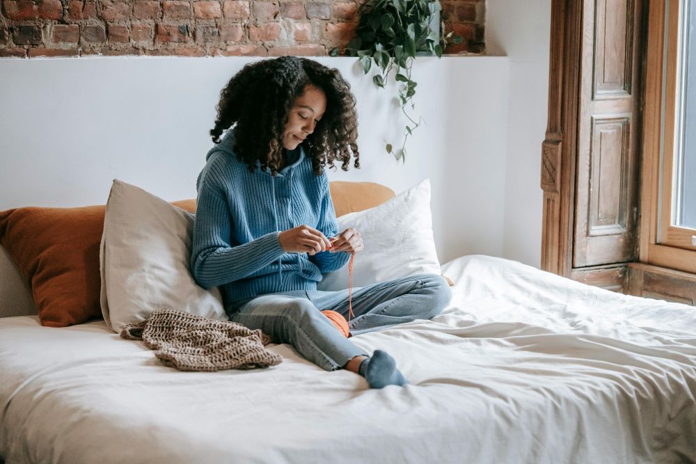 A woman sits in a comfy, minimalist bed with natural colours in a soft sweater while knitting