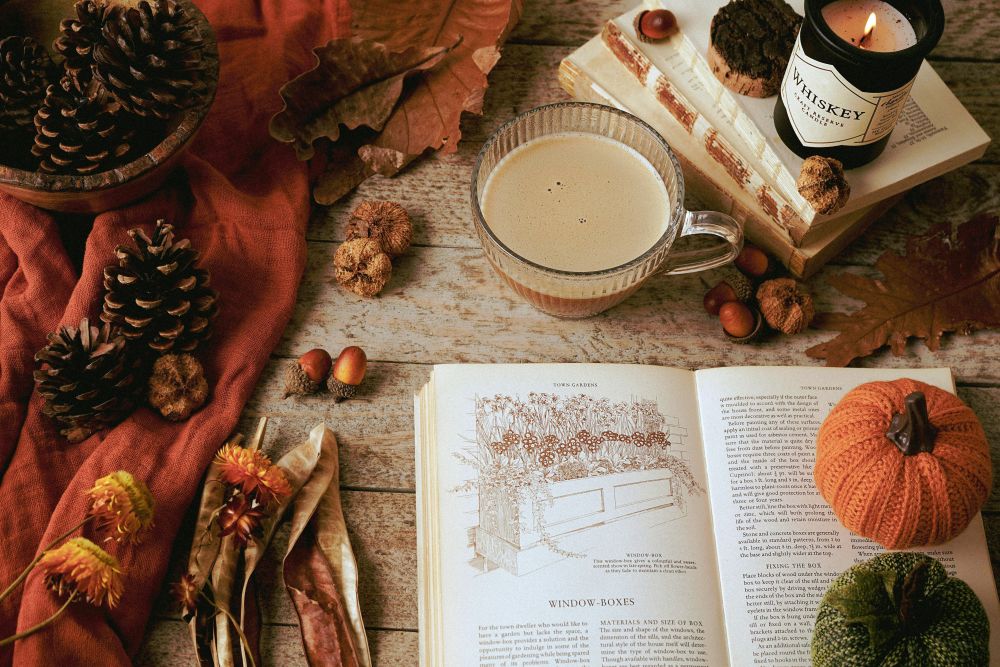 A cosy autumn vignette features a mug of coffee, a lit candle, a few books, a blanket, pinecones, and other earthy decor